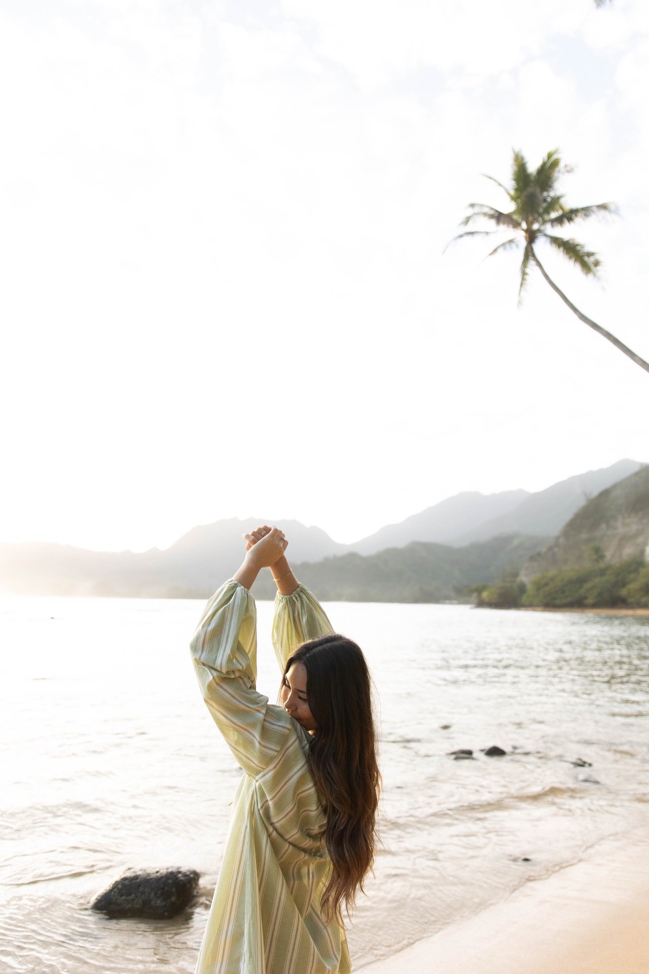 Woman in a green striped dress on the beach with a palm tree and Kauaʻi mountains behind