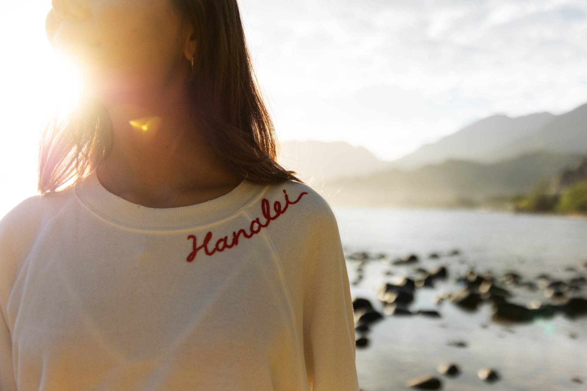 Woman in a Hanalei embroidered sweatshirt with sun flare over Hanalei Bay