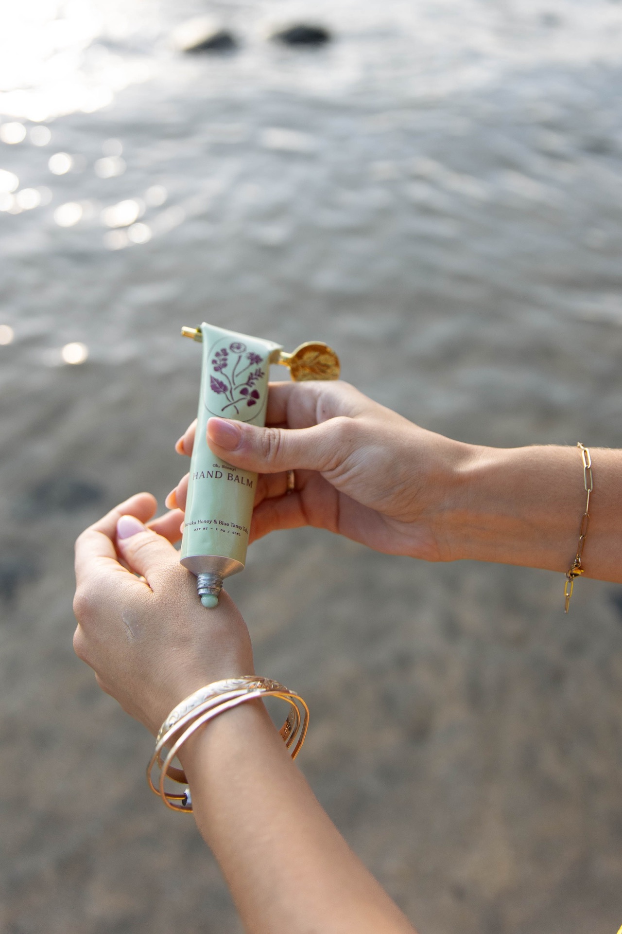 Hands applying My Neighbors hand balm by the shoreline with bracelets