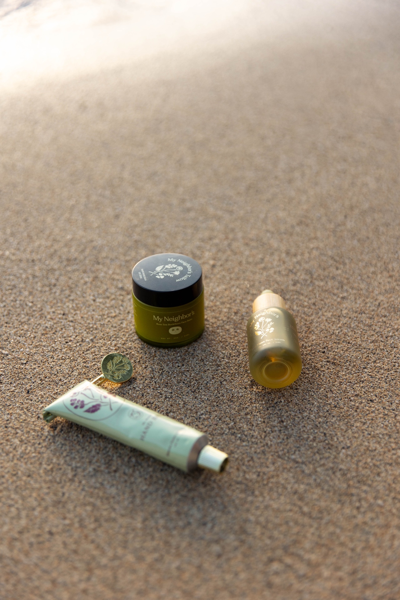 My Neighbors balm, hand cream and serum arranged on golden Kauaʻi sand