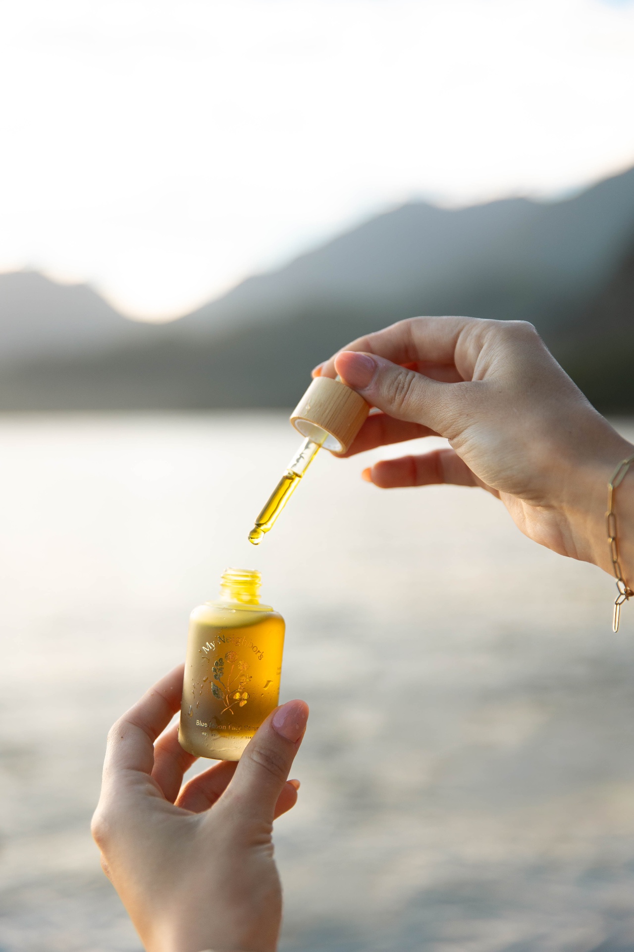 Hands holding a My Neighbors Blue Moon face serum and dropper above Hanalei Bay