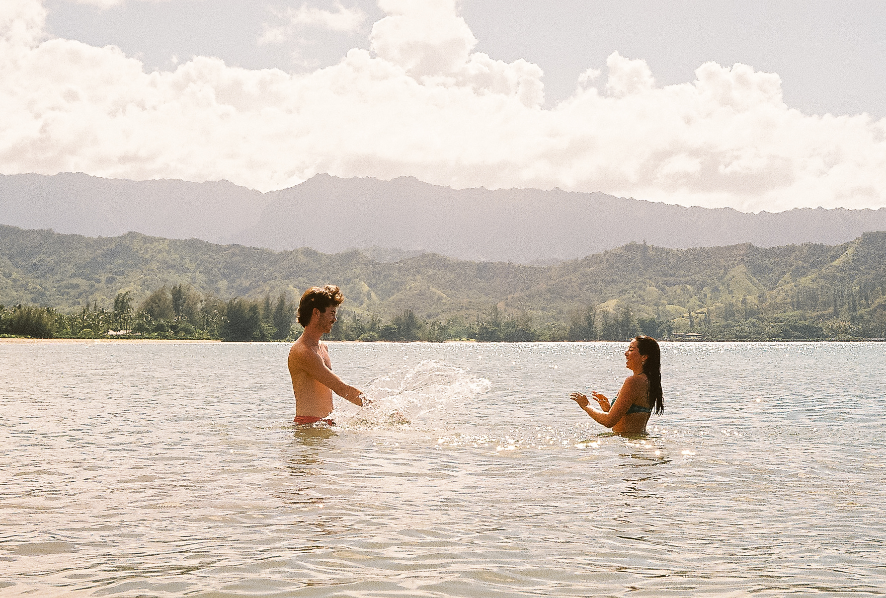 Couple playfully splashing each other in Hanalei Bay with green mountains in the distance, Kauaʻi