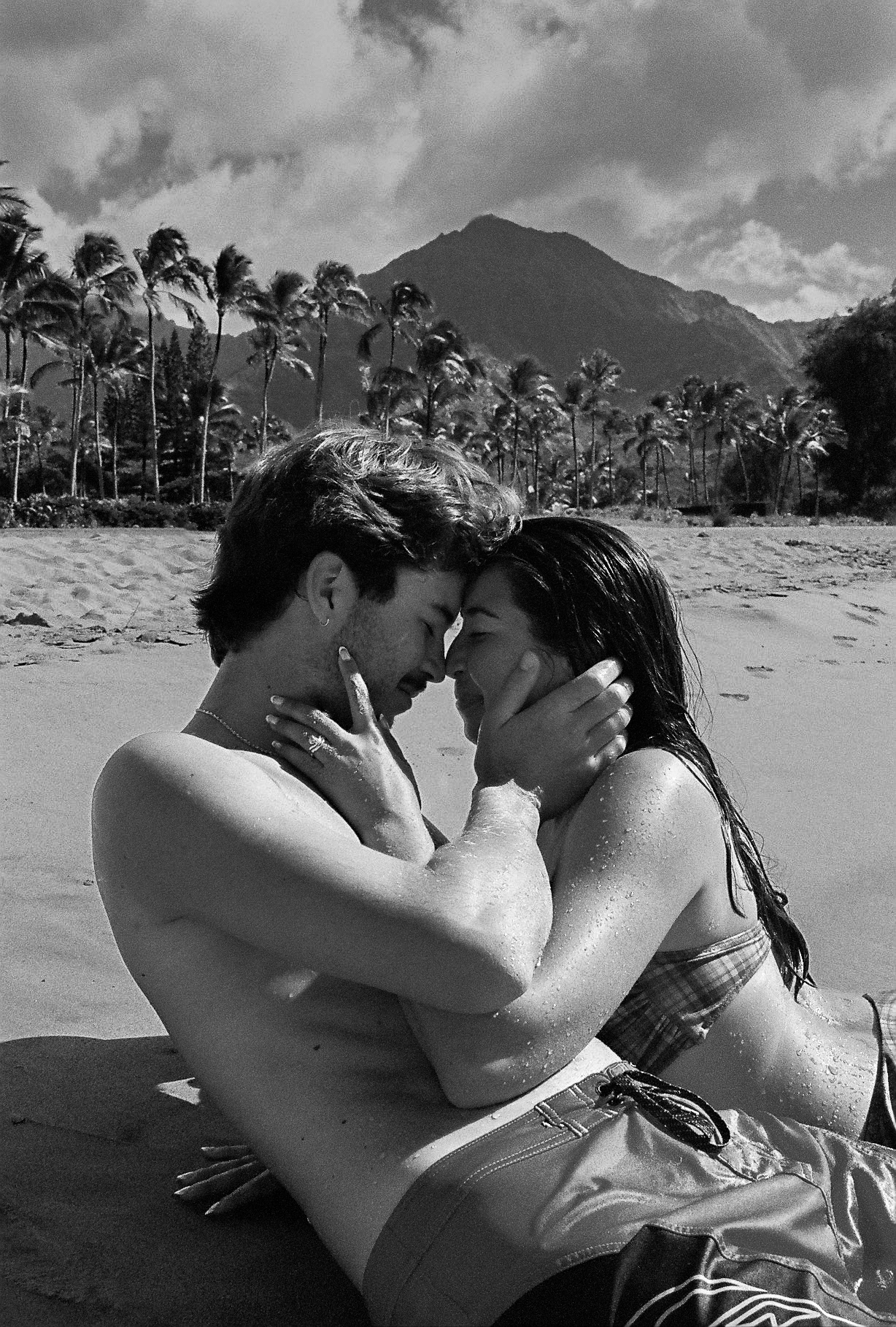 Black and white photo of couple touching foreheads on the beach with palms and mountains behind, Kauaʻi