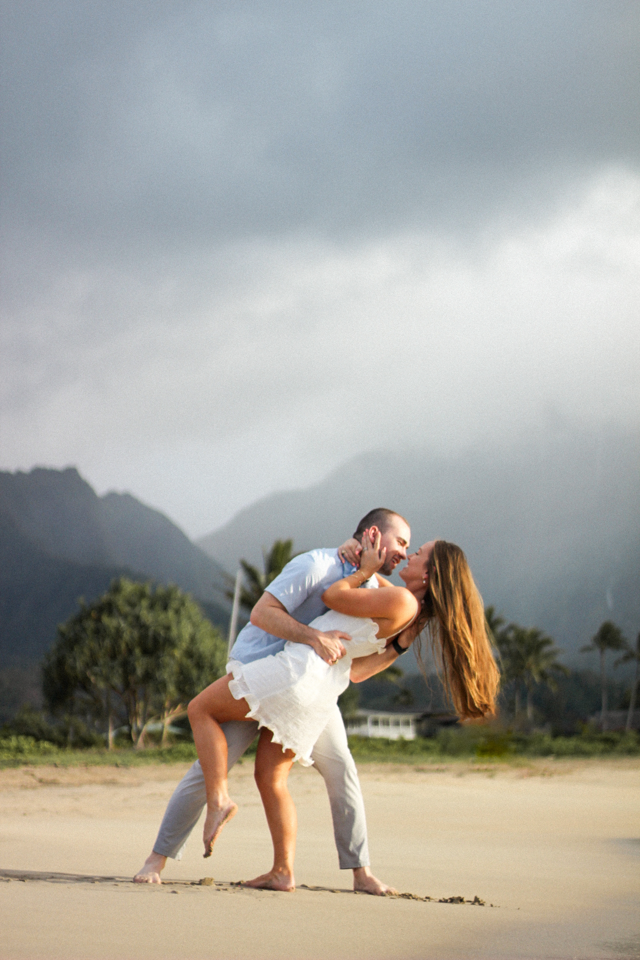 Couple in a dipped kiss on the beach with dramatic mountains and stormy sky behind, Kauaʻi