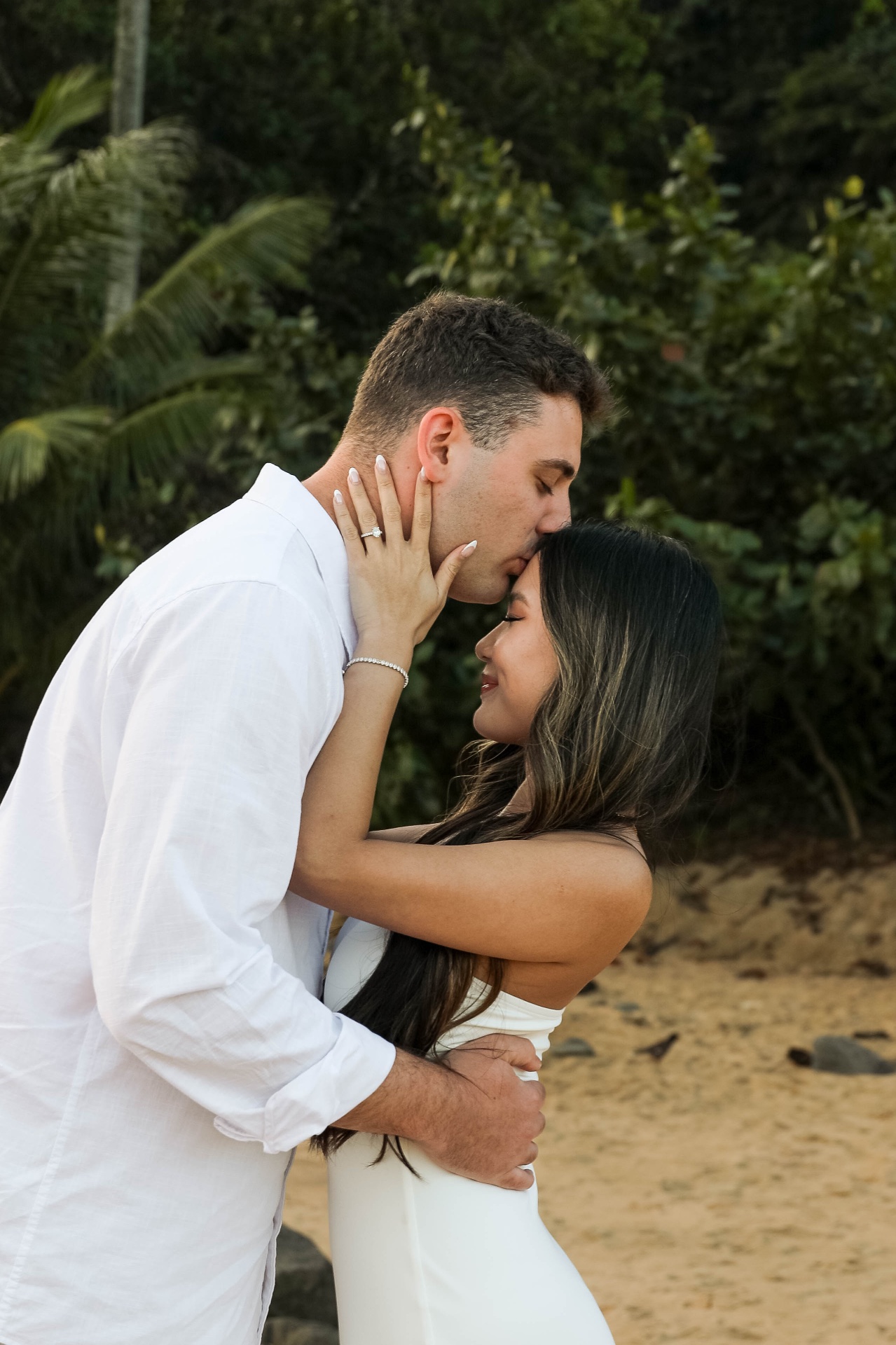 Groom kissing bride's forehead as she holds his cheek showing engagement ring, Kauaʻi