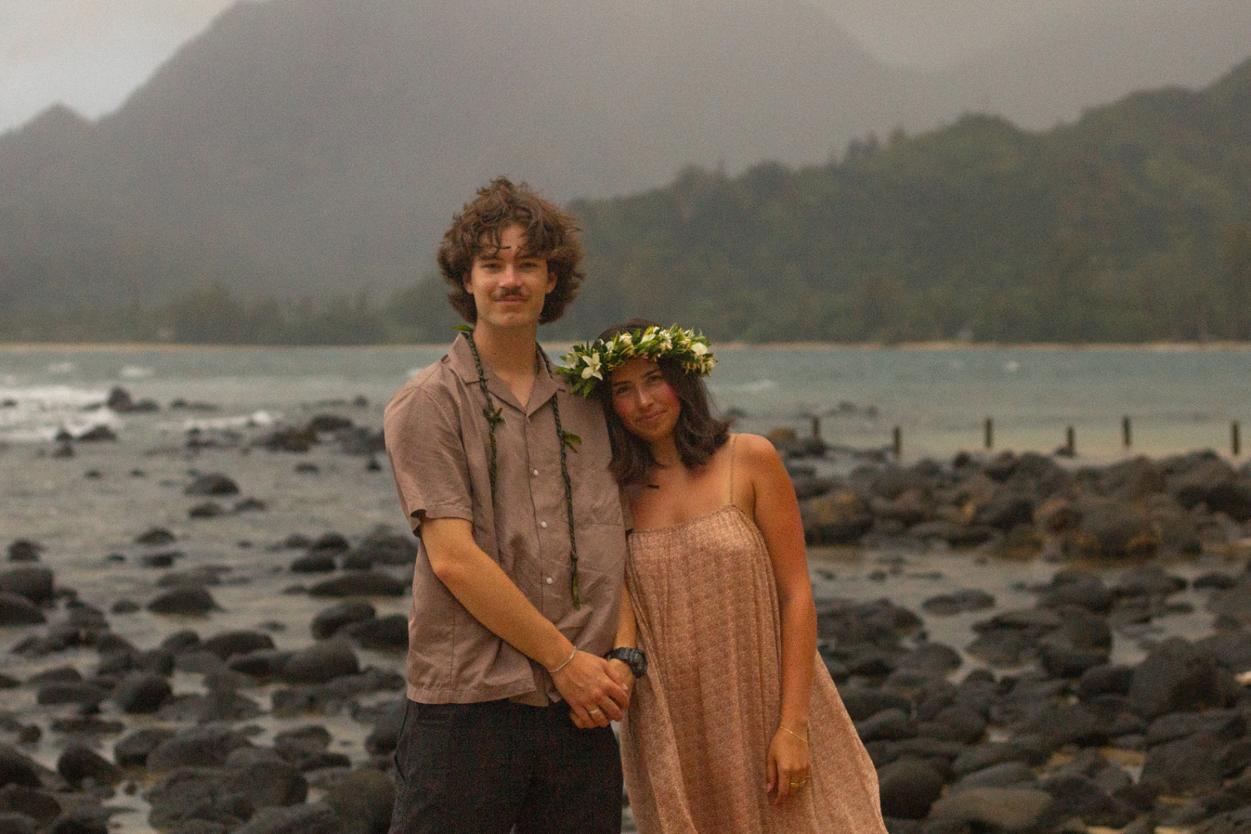 Couple in lei and haku flower crown holding hands on rocky shoreline with misty mountains, Kauaʻi