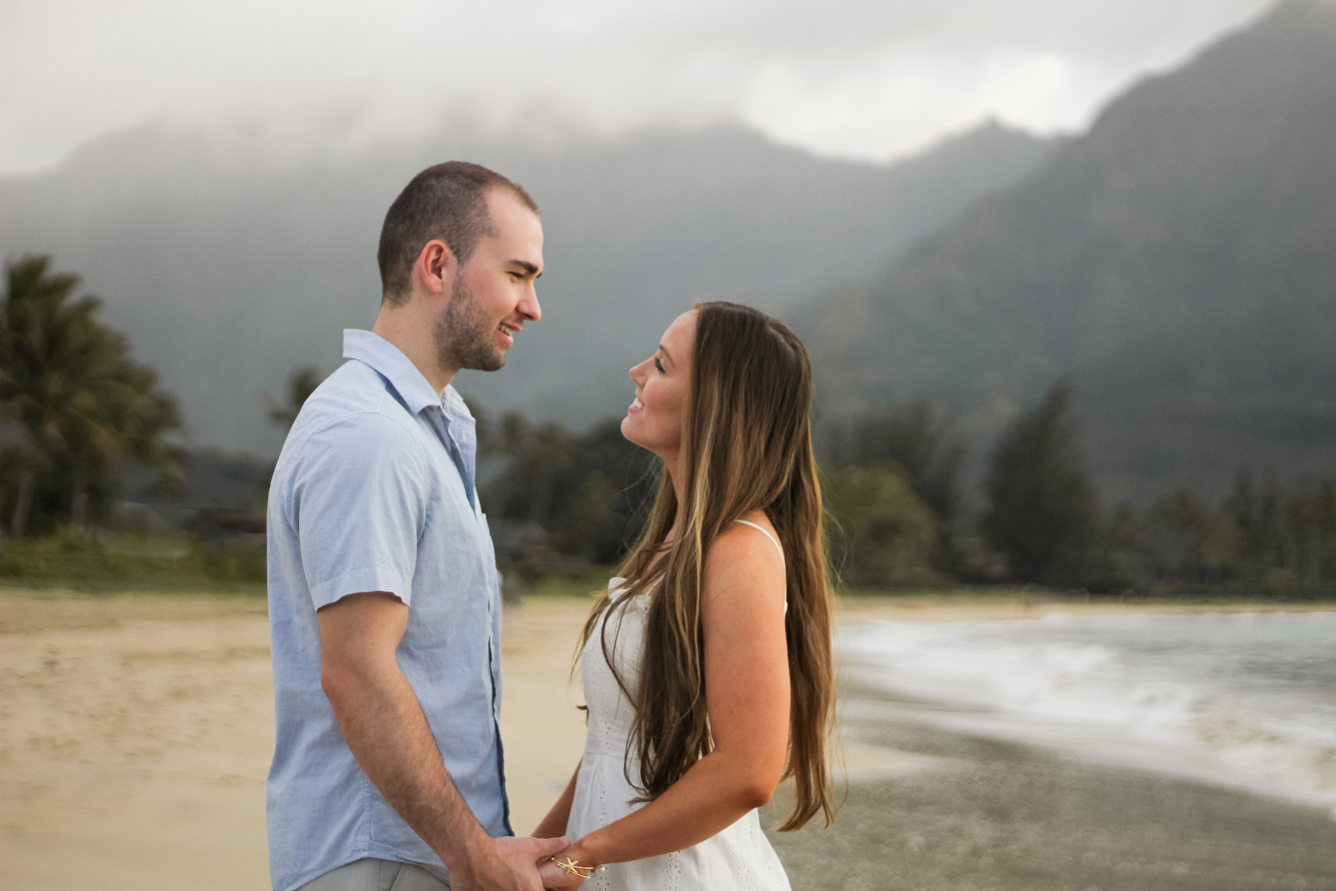 Couple smiling at each other and holding hands on the beach with misty Kauaʻi mountains behind