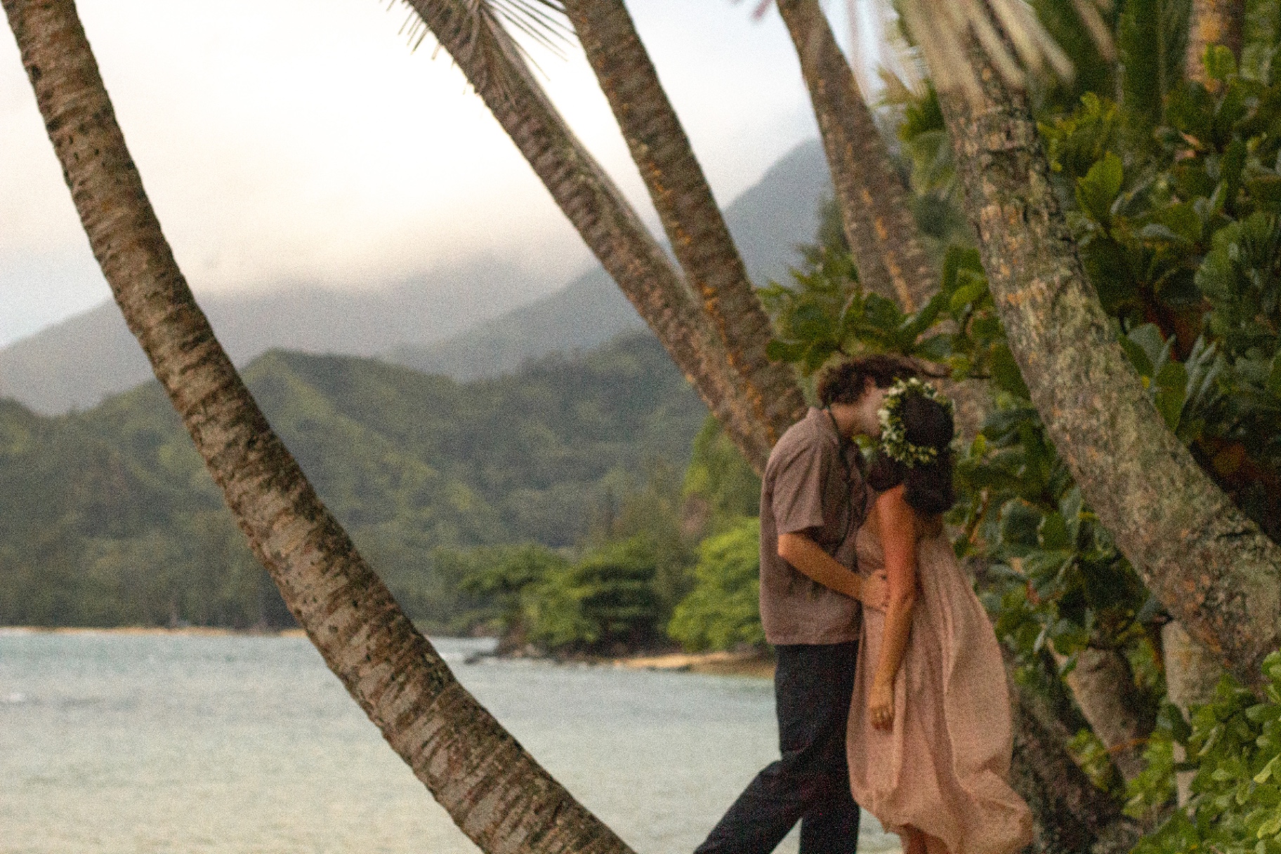 Couple kissing between palm trees with bay and mountains behind at dusk, Kauaʻi