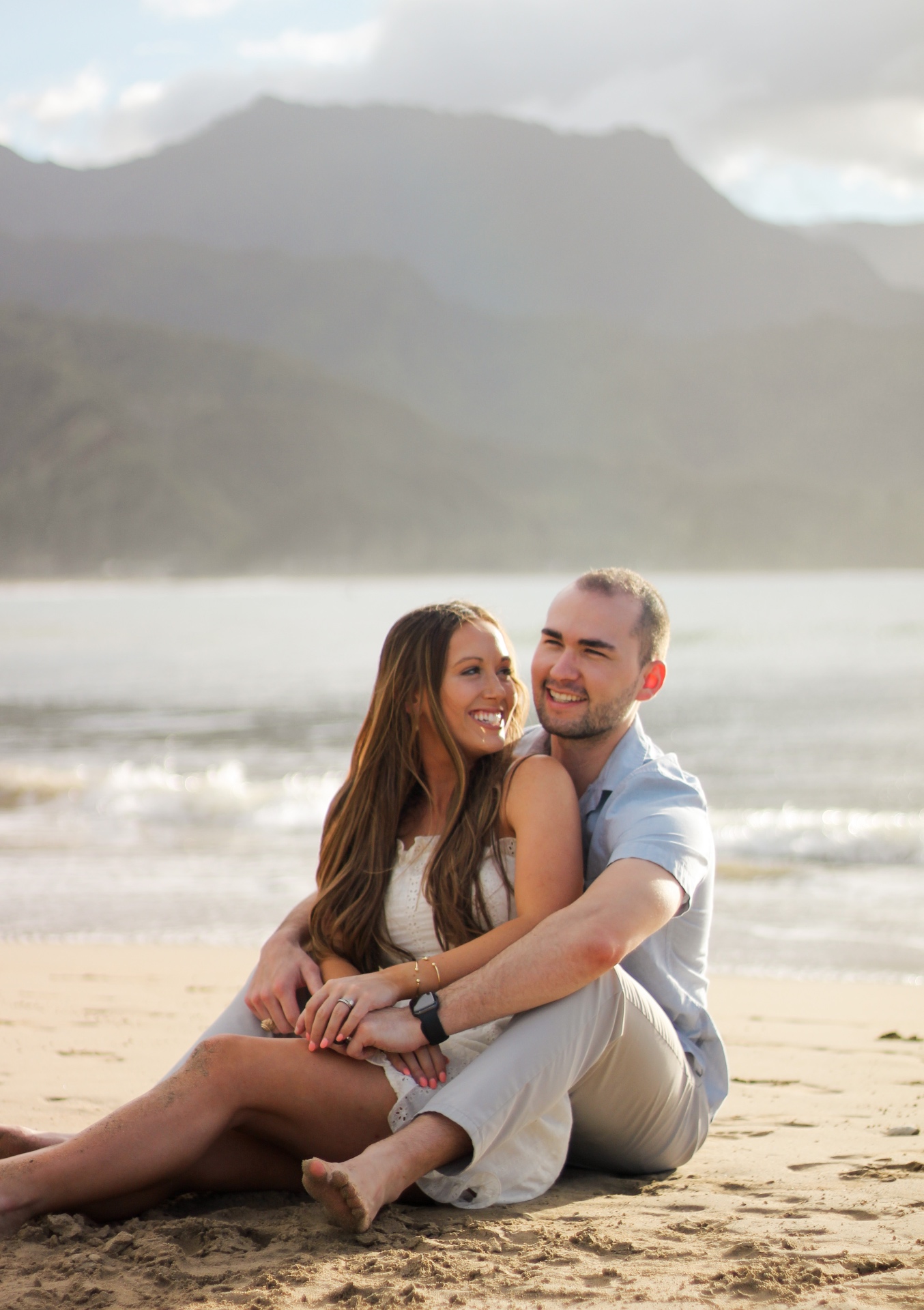 Couple sitting on the sand laughing together with Hanalei Bay and mountains behind, Kauaʻi