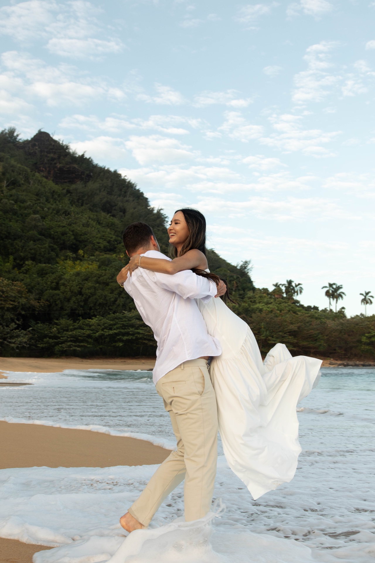 Groom lifting smiling bride in a white dress at the shoreline with green mountains behind, Kauaʻi
