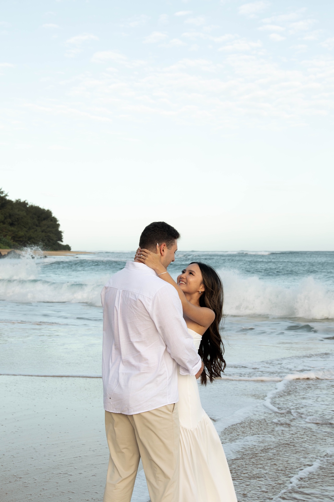 Bride smiling up at groom while embracing on the shoreline as waves crash behind, Kauaʻi