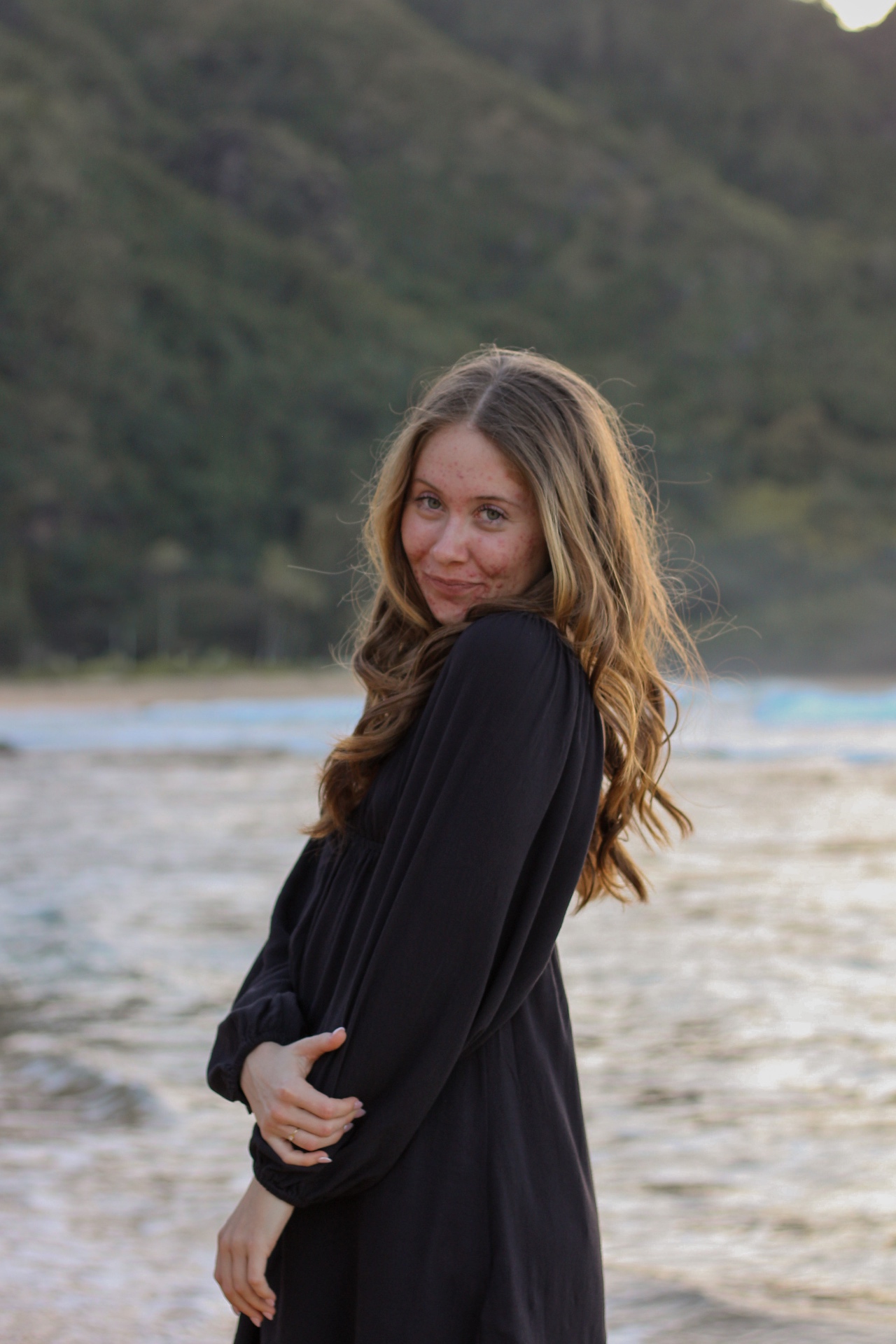 Portrait in flowing black dress at the shoreline with green mountains behind, Kauaʻi