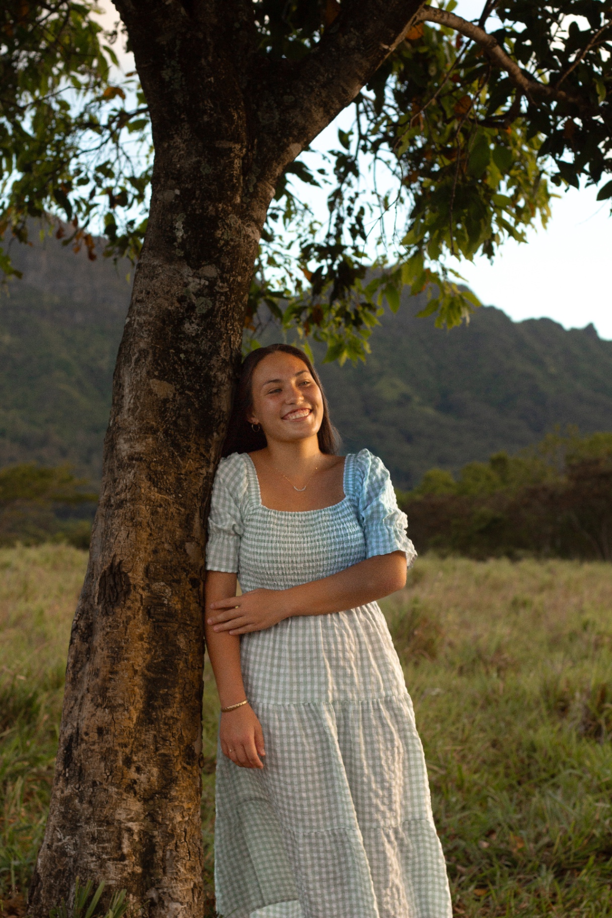 Portrait leaning against a tree in a gingham dress with mountains behind, Kauaʻi